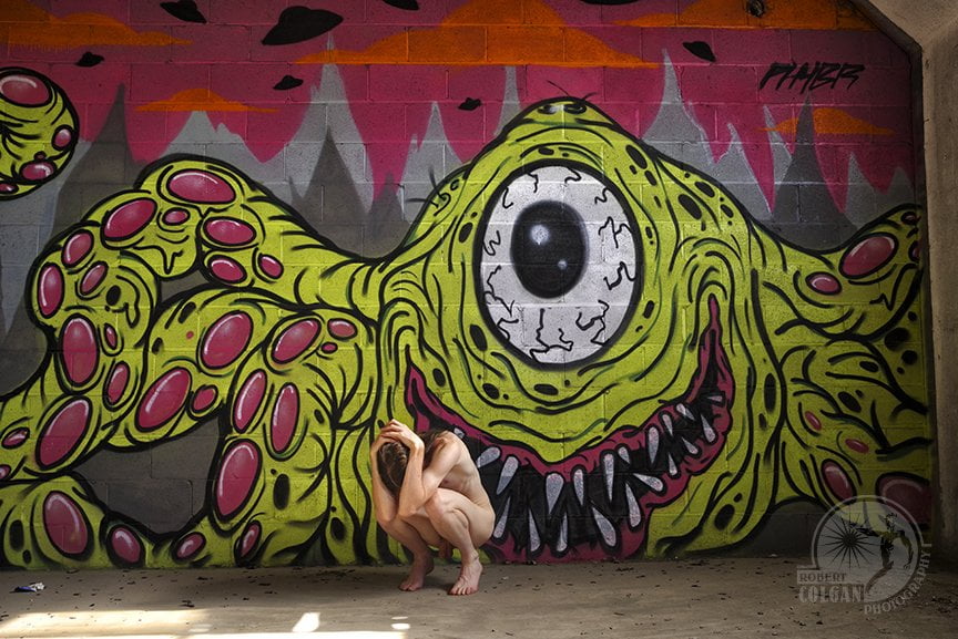 Nude man in fetal position in front of graffiti wall with one-eye's monster painted on it