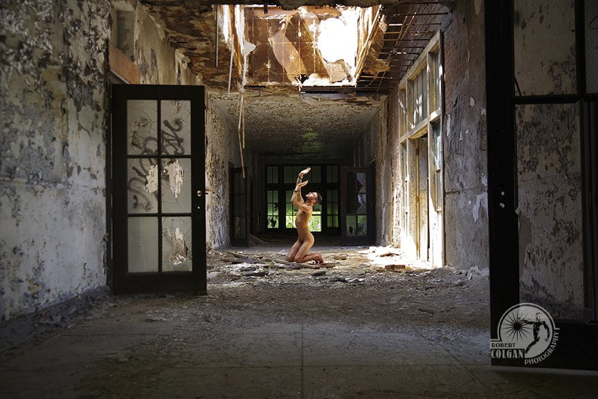 nude man handcuffed and kneeling in demolished hallway looking toward skylight