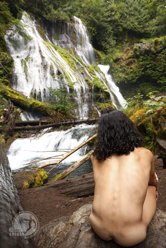 nude man sits watching waterfall in woods