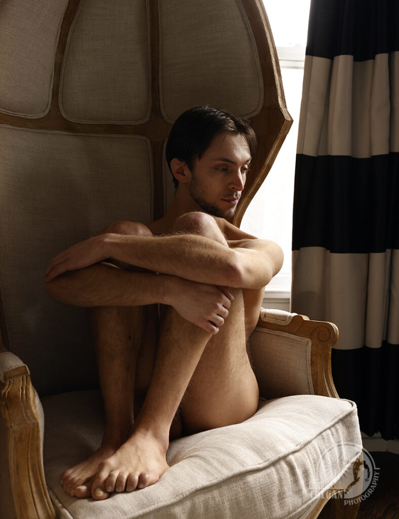 nude man curls up in chair by window