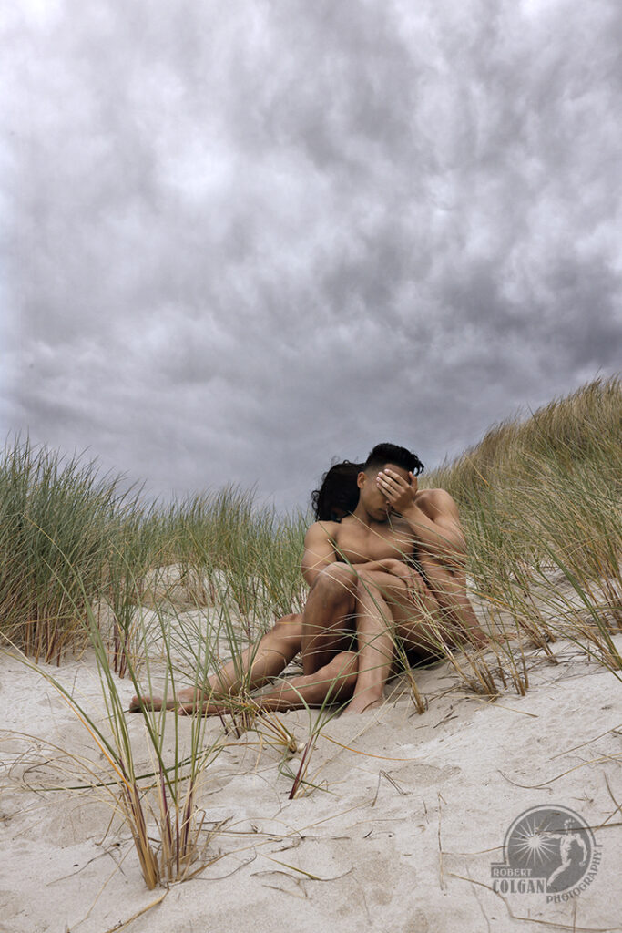 two nude men hide in the dunes, one covers the eyes of the other