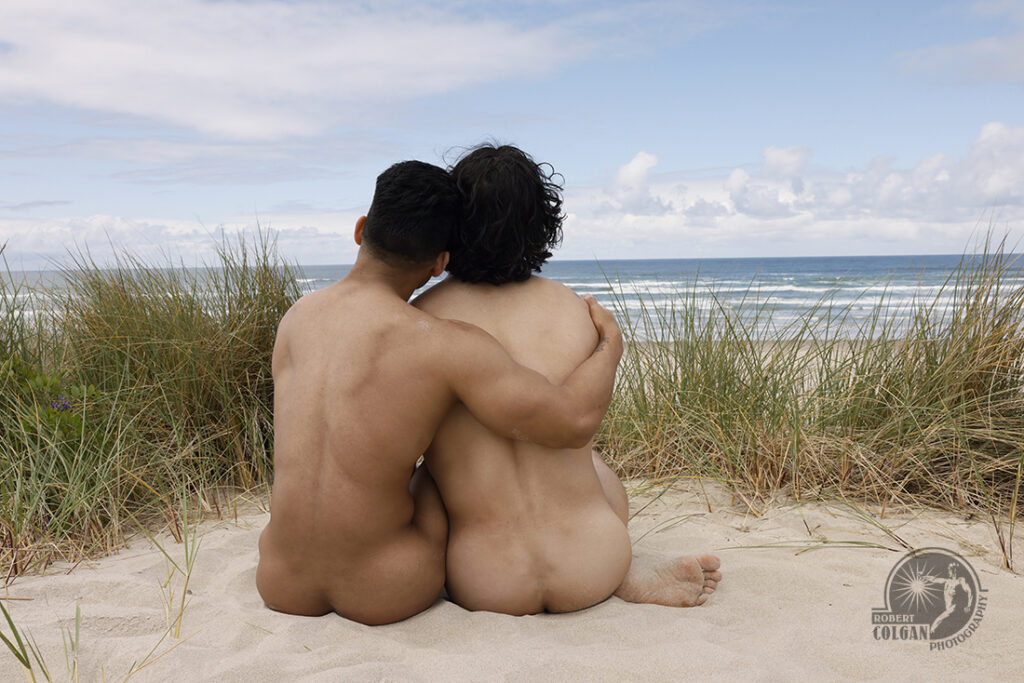 two nude men, photographed from behind, sit in sand dunes looking out at the ocean