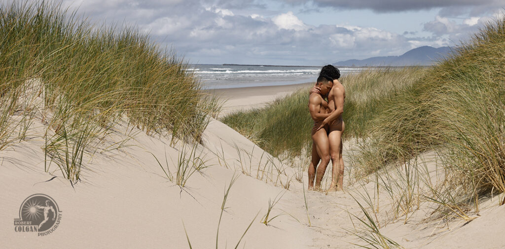 two nude men embrace in a hidden spot in the dunes