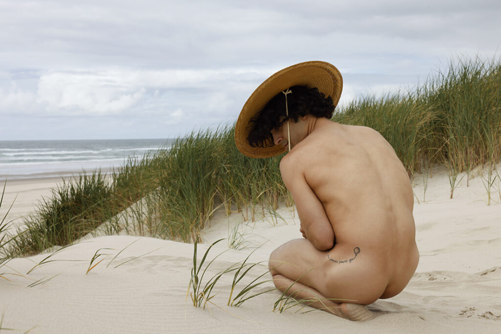 nude man curled up on beach with straw hat