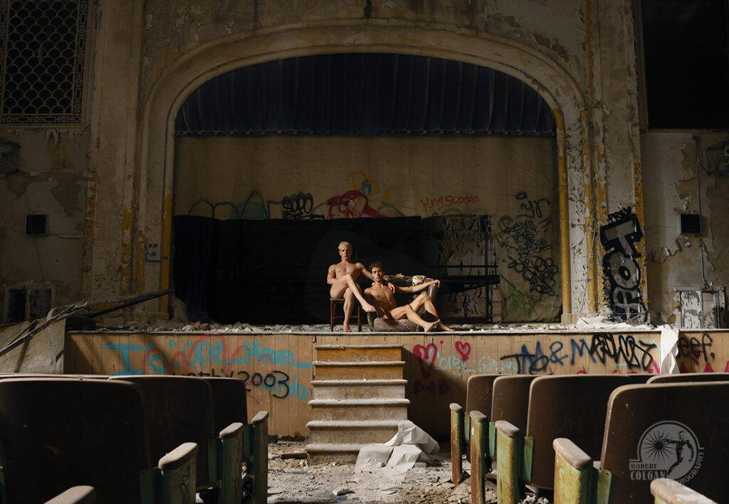 Two nude men sit stoically on a demolished school stage