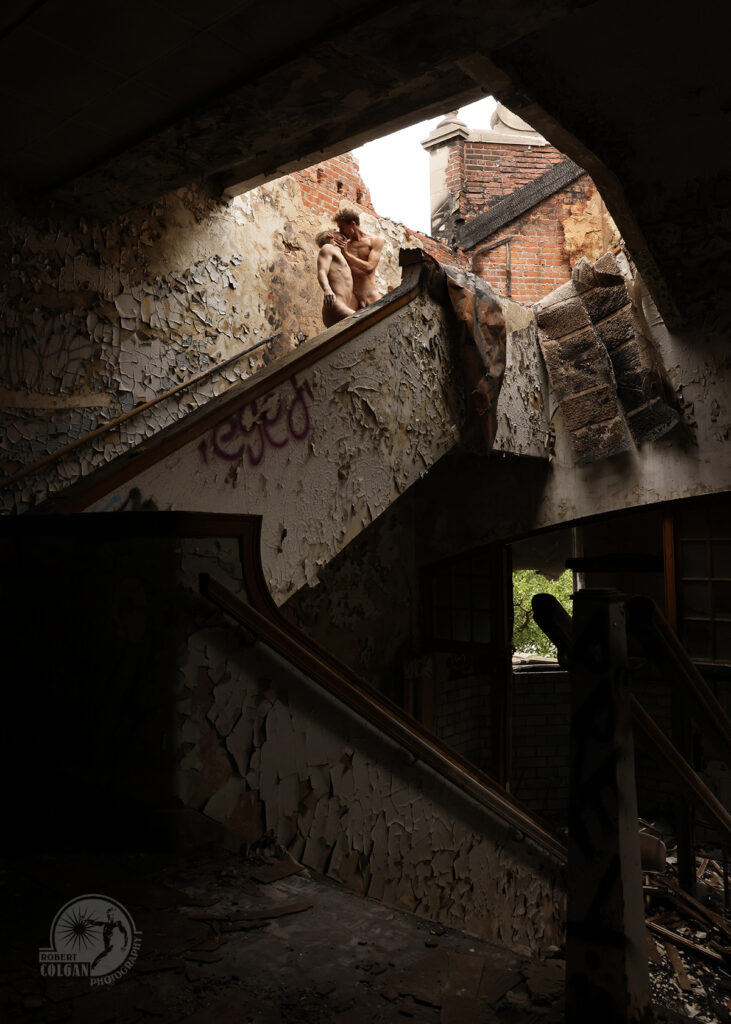 two nude men kiss at the top of stairs in a burned out building