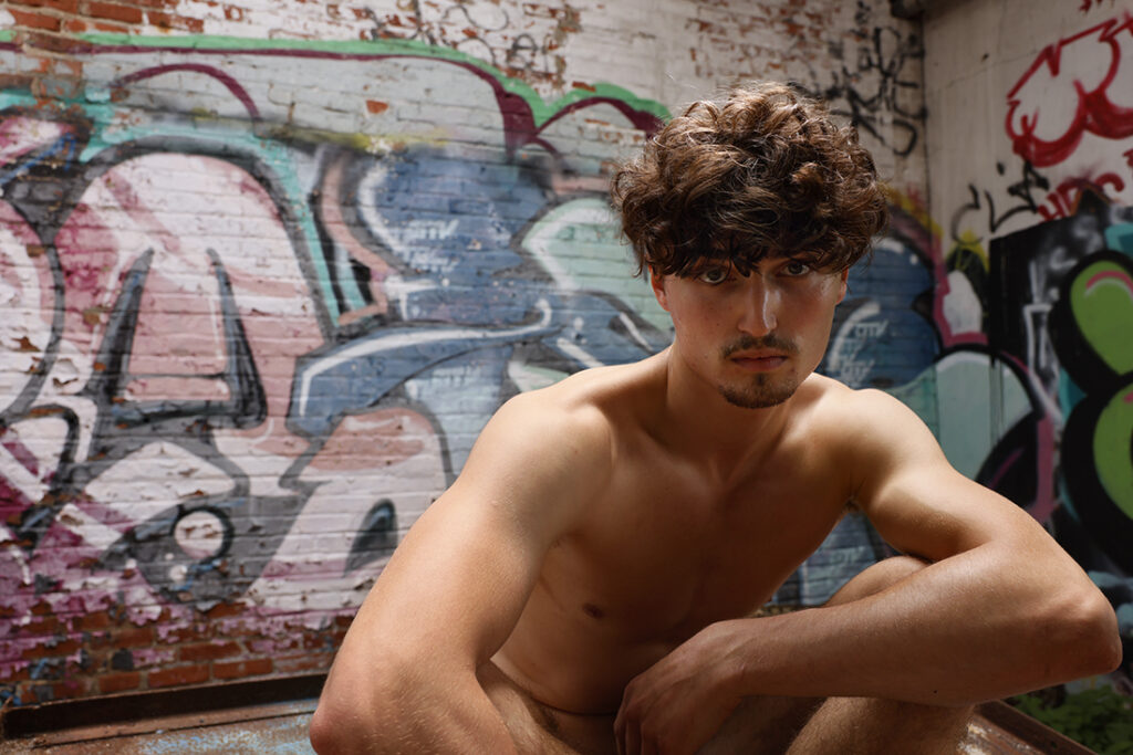 shirtless man staring at camera with graffiti behind