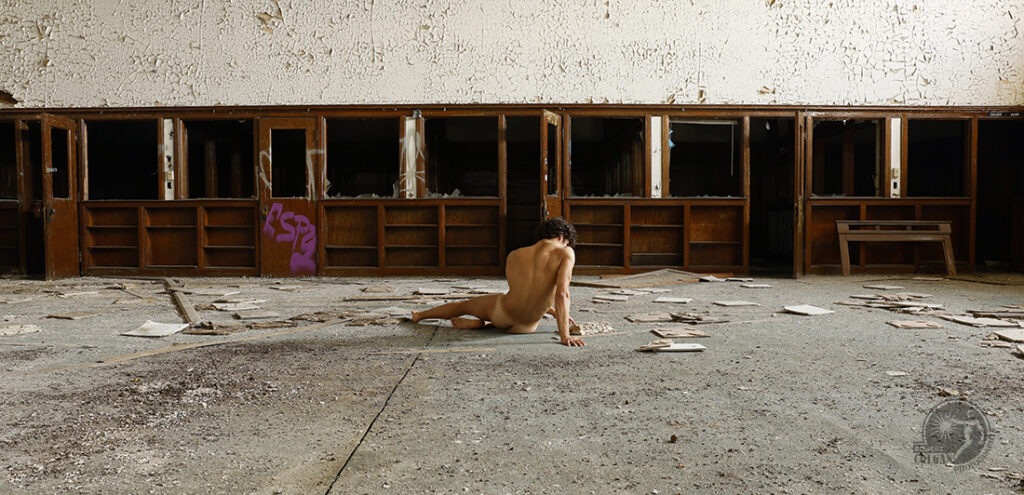 Nude man sits on the floor of an abandoned library looking at a rotting book