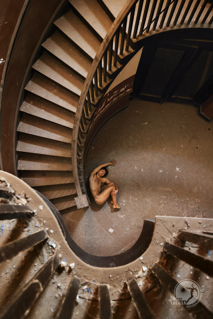 Nude man curled up at base of winding staircase