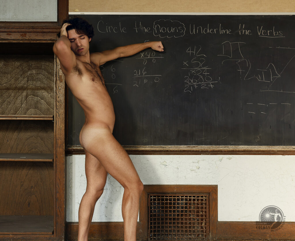 Nude man stands beside chalk board of abandoned school emphasizing text written on it