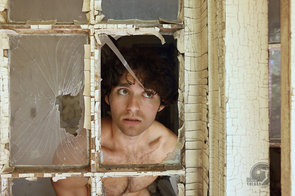 Shirtless man looks out through broken window with shard of glass pointing toward his eye